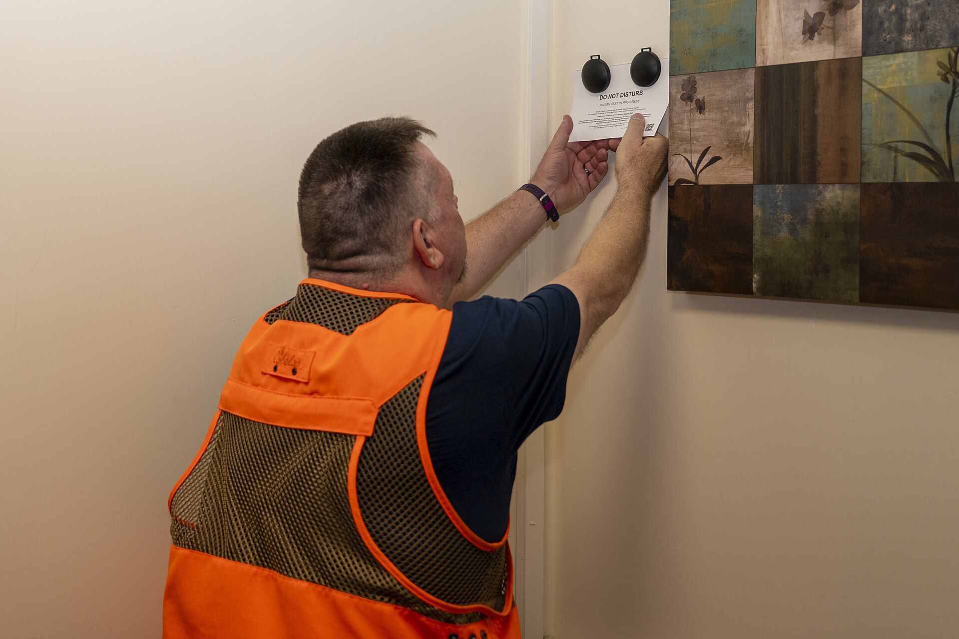 A worker in a safety vest picking up a radon detector from a wall.